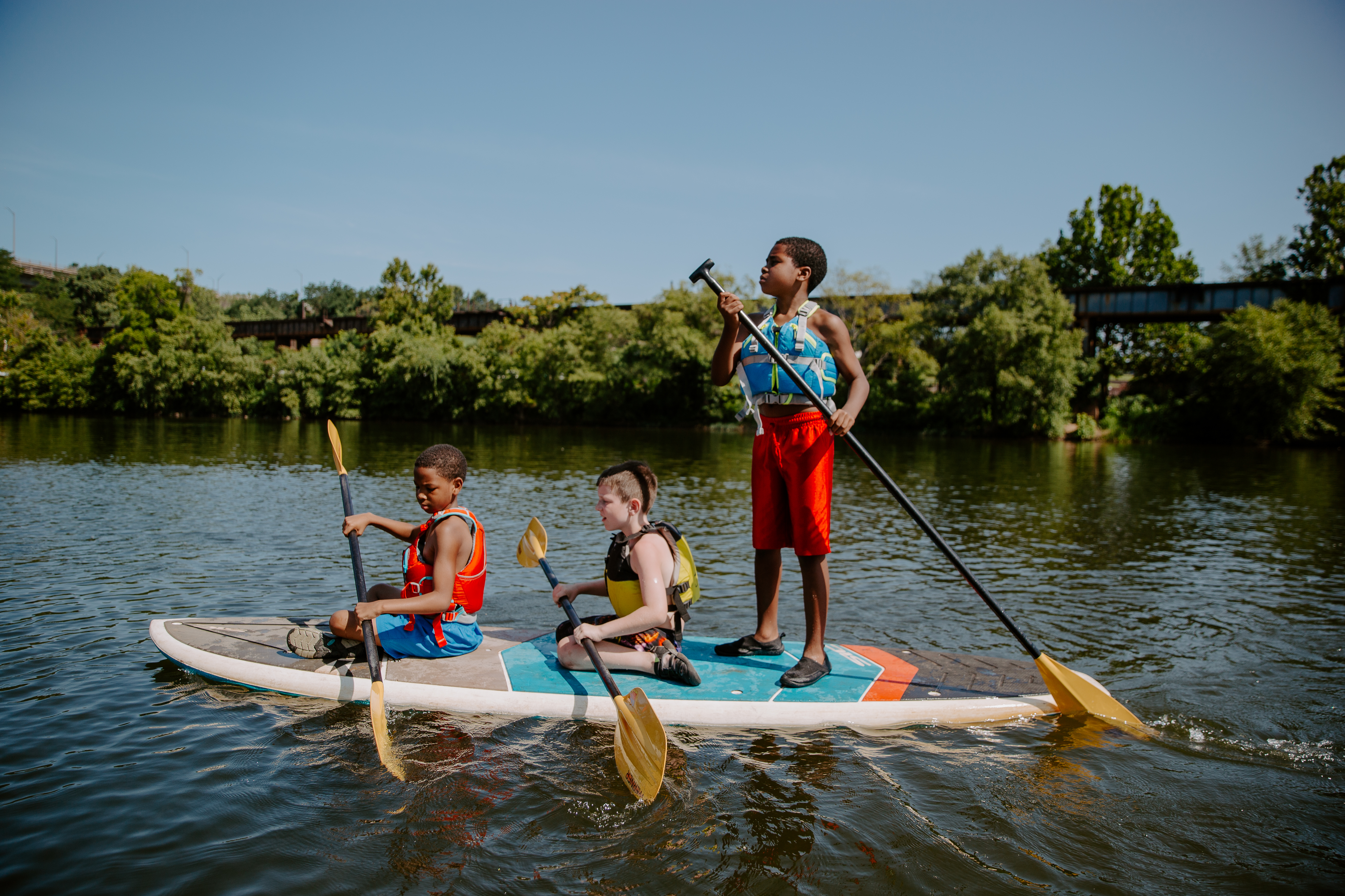 Children paddleboarding on the James River during an outdoor recreation program, wearing life jackets and using paddles on a sunny day